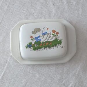 Può includere: Un burriera in ceramica bianca con coperchio rettangolare caratterizzato da una colorata illustrazione di due oche che indossano fiocchi blu a pois. Le oche sono posizionate su un prato verde con piccoli fiori arancioni e bianchi e una nuvola.