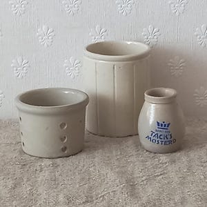 Op de afbeelding: Drie off-white keramische keukenhouders: een kleine kom met perforaties aan de zijkant, een hogere cilindrische houder met verticale ribbels en een klein rond potje met een blauw kroonlogo en de tekst TACK'S MOSTERD.