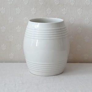 Pode incluir: Um vaso de cerâmica branca com formato de barril, apresentando um acabamento liso e brilhante com detalhes horizontais em relevo nas seções superior e inferior. O recipiente possui uma abertura larga e um corpo arredondado que se estreita.