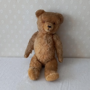 Puede incluir: Un oso de peluche vintage marrón con ojos de botón y una nariz cosida, sentado sobre una superficie blanca.