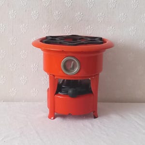 Peut inclure: Un réchaud de camping portable orange vif doté d'une grille métallique noire sur le dessus, d'une base de réservoir de carburant circulaire et d'une petite fenêtre d'observation ronde en verre à l'avant, avec un bouton de commande noir.