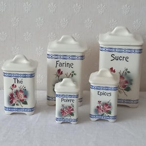 Pode incluir: Um conjunto de cinco potes de cozinha em cerâmica branca com tampas, apresentando bordas decorativas azuis e padrões florais. Cada pote está rotulado em preto com as palavras Thé, Farine, Poivre, Epices e Sucre, indicando o conteúdo.