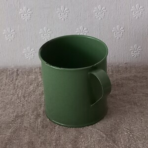 Può includere: Una tazza in smalto verde con manico curvo. La tazza è di un colore verde pieno e smorzato. La tazza è cilindrica con un bordo leggermente angolato. Lo sfondo è un muro strutturato bianco sporco con un motivo floreale.