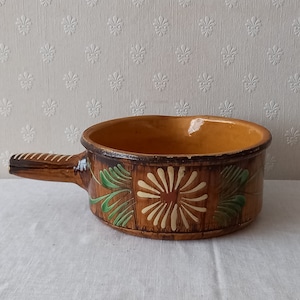 May include: A brown ceramic pot with a handle, decorated with green and white floral designs. The pot has a glazed interior.