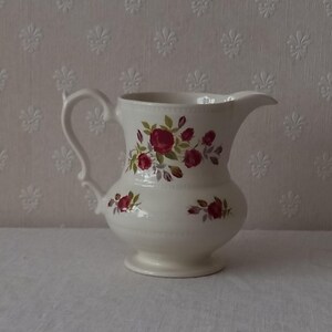 May include: White ceramic pitcher with a floral design featuring red roses and green leaves. The pitcher has a handle and a spout.