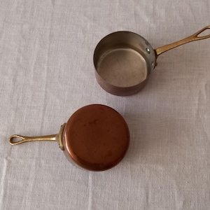 Puede incluir: Dos pequeñas cacerolas de cobre con asas de latón. Una cacerola está abierta, revelando el interior, mientras que la otra está invertida, mostrando la base de cobre. Las asas tienen un lazo para colgar. Sobre una superficie de color neutro.