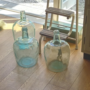 Op de afbeelding: Drie grote, heldere glazen mandflessen met ronde lichamen en smalle halzen. Elk vat heeft een licht blauwgroene tint en een glad, reflecterend oppervlak. De containers zijn in een groep gerangschikt en tonen hun transparante, bolvormige vormen en stevige glazen constructie.