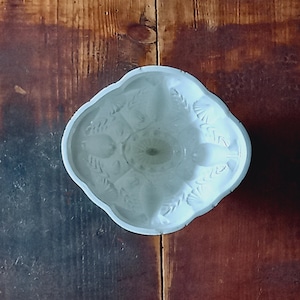 May include: A white, oval-shaped ceramic mold featuring a detailed, embossed pattern of wheat stalks and shell motifs. The interior surface is smooth and white, with a scalloped edge design that follows the contour of the oval shape.