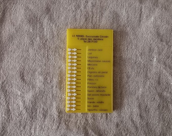 May include: A yellow plastic card with a list of French grocery items, including "Jambon-lard" and "Lait." The card also has the text "LE MANS - Succursale Citroen" and a phone number. The card is on a textured, neutral background.