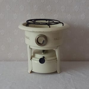 Op de afbeelding: Off-white vintage tafelkachel met een zwart metalen rooster. De kachel heeft een cirkelvormig ontwerp met de woorden "ORIGINAL HALLE" aan de zijkant gedrukt. Het heeft een cirkelvormige opening en een zwarte knop.