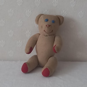 Puede incluir: Un oso de peluche hecho a mano de color beige, con detalles rojos en las manos y los pies. El oso tiene ojos azules de botón, una sonrisa cosida y una nariz roja. Está sentado sobre una superficie blanca.