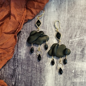 May include: A pair of black cloud-shaped earrings with dangling black beads. The earrings are made of polymer clay and have a gold-tone finish.