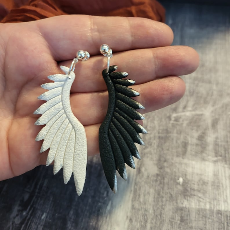 Pu&ograve; includere: Un paio di orecchini pendenti con un design ad ala d'angelo in bianco e nero. Le ali sono realizzate in materiale testurizzato e hanno una finitura argentata.