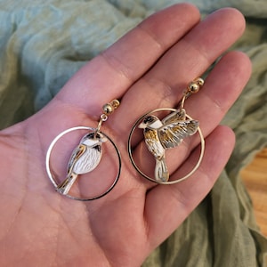 May include: A pair of gold hoop earrings with a white and gold bird charm hanging from each hoop.