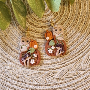 May include: Handmade earrings with a seasonal autumn theme. Each earring features a brown pumpkin base, an owl, a small orange pumpkin, mushrooms, and white flowers. The owl is tan with white polka dots. The earrings are on gold-coloured hooks.