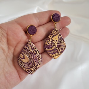 May include: A pair of dangle earrings with a purple base and gold patterned design. The earrings are shaped like a diamond.
