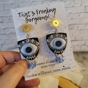 May include: A pair of handmade earrings featuring a blue evil eye design with black and silver accents. The earrings are made of polymer clay and have gold-toned accents. The earrings are hanging from a gold-toned chain with a sunburst charm.