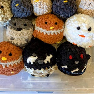 May include: A collection of handmade, crocheted chick-shaped toys in various colors. The chicks have black eyes, yellow beaks, and some have white or gray accents. The toys are soft and fluffy, with a whimsical design.
