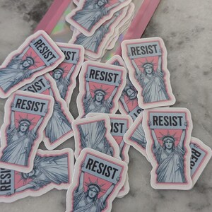 May include: A collection of stickers featuring the Statue of Liberty holding a sign that reads "RESIST". The stickers have a pink and blue color scheme with a white border. Some stickers are in a pink holographic bag.