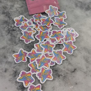 May include: A collection of star-shaped stickers with a rainbow gradient design and the word "Resist" in a script font. The stickers are scattered on a light-colored surface, with a pink pouch in the background.