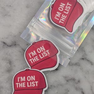 May include: Red baseball cap stickers with the text "I'M ON THE LIST" in white. The stickers are in a clear, sealed bag. Several stickers are visible, with a few loose on a marble surface.