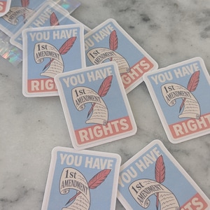 May include: A collection of stickers with a light blue background, featuring a scroll with a feather and the text "YOU HAVE 1st AMENDMENT RIGHTS" in red and white. The stickers are rectangular with rounded corners.