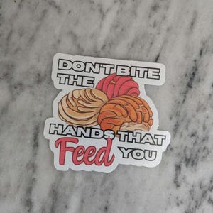 May include: A white sticker with the text "Don't Bite the Hands That Feed You" in black and red lettering. The sticker features a cartoon image of three colorful Mexican sweet breads.