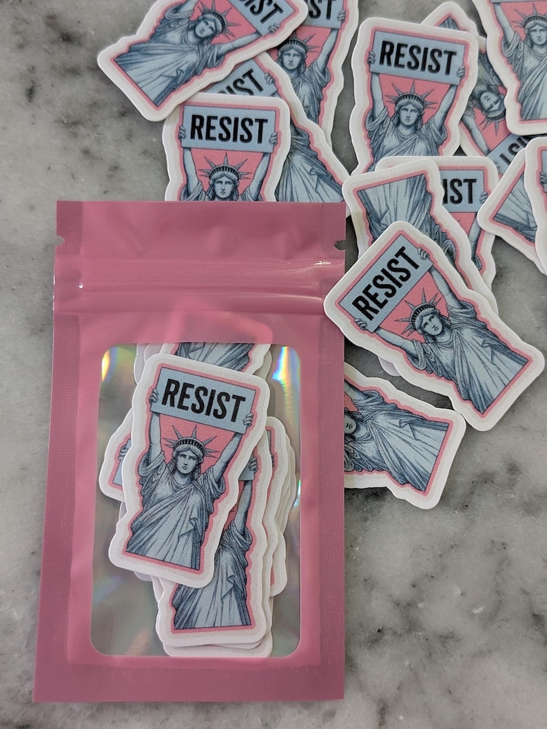 May include: A collection of stickers featuring the Statue of Liberty holding a sign that reads "RESIST". The stickers have a pink and blue color scheme with a white border. Some stickers are inside a pink, clear pouch.