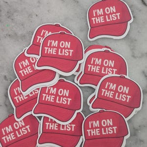 May include: A collection of red baseball cap stickers with the text "I'M ON THE LIST" in white. The stickers are scattered on a light gray surface. The caps have a classic design.