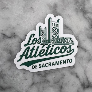 May include: A white sticker with a green graphic of a bridge and the words "Los Atléticos DE SACRAMENTO" in green. The sticker has a white border and is on a gray surface.