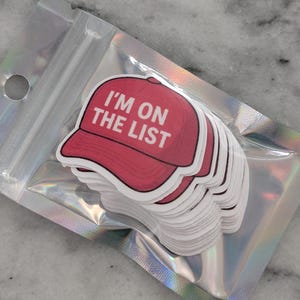 May include: A stack of stickers featuring a red baseball cap design with the text "I'M ON THE LIST" in white. The stickers are packaged in a clear, iridescent pouch.