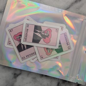 May include: A collection of pink and white stickers featuring a clown with a red nose and a pink afro, wearing a black suit and tie. The stickers are labeled "EL PENDEJO" and numbered "47". They are inside a holographic, iridescent plastic bag.