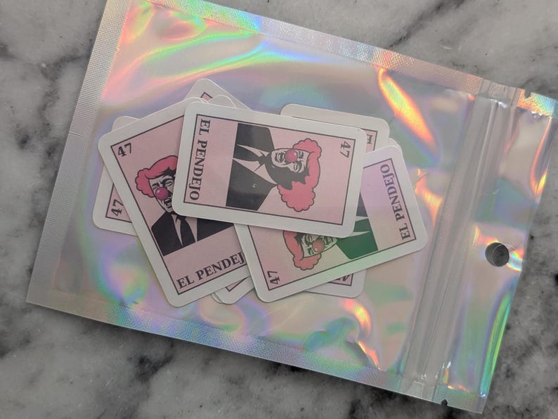 May include: A collection of pink and white stickers featuring a clown with a red nose and a pink afro, wearing a black suit and tie. The stickers are labeled "EL PENDEJO" and numbered "47". They are inside a holographic, iridescent plastic bag.