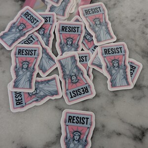 May include: A collection of stickers featuring the Statue of Liberty holding a sign that reads "RESIST." The stickers have a pink background with a light blue border and the statue is in light blue. The stickers are scattered on a marble surface.