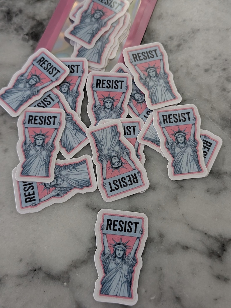 May include: A collection of stickers featuring the Statue of Liberty holding a sign that reads "RESIST." The stickers have a pink background with a light blue border and the statue is in light blue. The stickers are scattered on a marble surface.