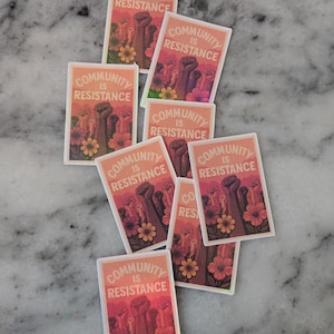 May include: A collection of stickers with the text "COMMUNITY IS RESISTANCE". The stickers feature a design of raised fists, flowers, and a gradient background of orange and pink.