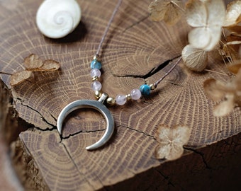 May include: A silver crescent moon pendant necklace with a string of blue, pink, and gold beads. The necklace is displayed on a rustic wooden surface, with a seashell and dried flowers in the background.