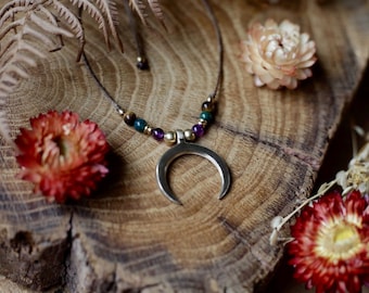 Puede incluir: Collar con colgante de media luna plateada con cuentas de piedras preciosas de colores. El collar descansa sobre una superficie de madera con flores secas. Un accesorio de estilo bohemio.