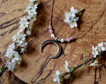 May include: A silver crescent moon pendant necklace with pink and white beads on a brown cord. The necklace is displayed on a wooden surface with white flowers.