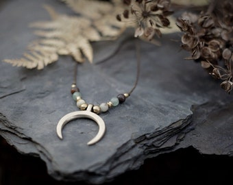 Puede incluir: Un collar con un colgante de media luna de plata, un cordón de cuero marrón y cuentas verdes y marrones.