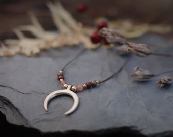 Puede incluir: Un collar con un colgante de media luna de plata, un cordón marrón y cuentas de piedras preciosas rosas y rojas.