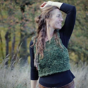 Könnte beinhalten: Eine grüne Strickweste mit strukturiertem Muster wird über einem schwarzen Langarmshirt getragen. Das Outfit umfasst einen braunen Ledergürtel mit quadratischer Schnalle und einen beigen Rock. Der Hintergrund zeigt hohes Gras und eine verschwommene Herbstlandschaft.