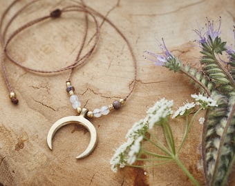 Puede incluir: Un collar con un colgante de media luna y un cordón marrón. El collar presenta pequeñas cuentas doradas y piedras preciosas de color claro. El collar se exhibe sobre una superficie de madera, con ramitas de flores blancas y moradas.