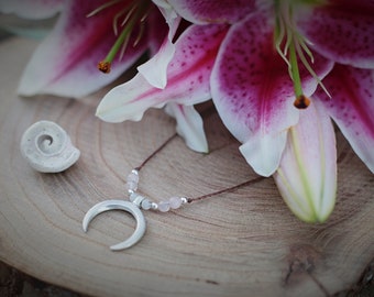 May include: A silver crescent moon pendant necklace with pink beads on a brown cord. The necklace is lying on a wooden surface with a white seashell and pink lilies.