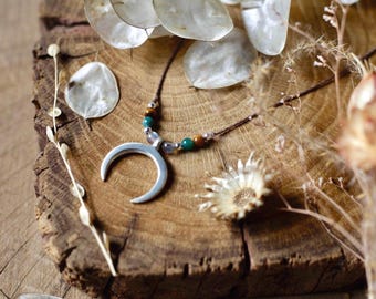 Puede incluir: Collar con colgante de media luna plateada con cordón marrón, adornado con cuentas verdes y marrones. El collar descansa sobre una superficie de madera rústica, con flores y hojas secas en el fondo.