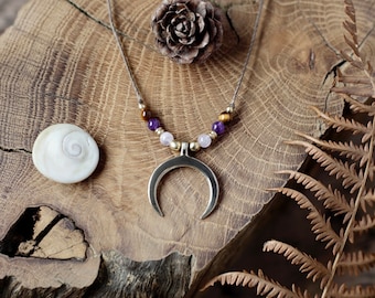 Puede incluir: Un collar con un colgante de media luna de plata con cuentas de piedras preciosas marrones, moradas y rosas en un cordón marrón. El collar se muestra sobre una superficie de madera con una concha y una piña.