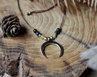 Puede incluir: Un collar de cordón de cuero marrón con un colgante de media luna y cuentas en tonos marrones, azules y verdes. El collar se muestra sobre una superficie de madera.