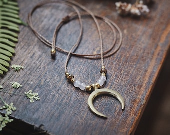 Puede incluir: Un collar con un colgante de media luna dorada, un cordón marrón y cuentas blancas y marrones.
