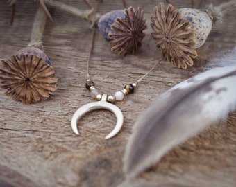 Puede incluir: Un collar con un colgante de media luna y cuentas rosas y marrones. El collar está ensartado en un cordón fino de color marrón claro. El fondo incluye vainas de semillas secas y una pluma sobre una superficie de madera.
