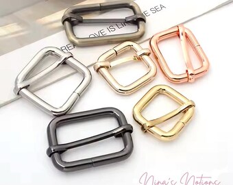 Op de afbeelding: Vijf metalen schuifgespen in verschillende afwerkingen: zilver, goud, zwart, rosé goud en antiek messing. De gespen zijn rechthoekig met een afgeronde rand en hebben een middelste stang die door de gesp schuift.
